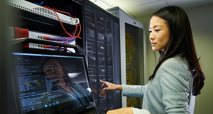 A young woman standing next to a rack of computer servers, looking at the screen.