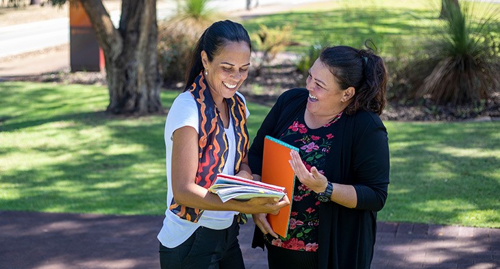 Two females smiling, holding books in a park setting.