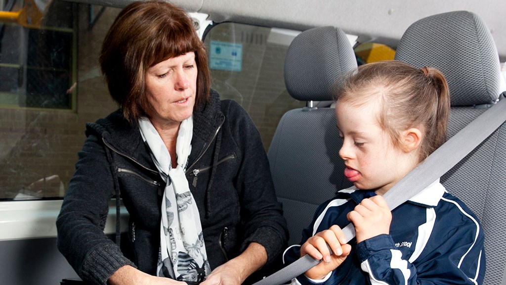 Female support officer helping young a female student with a disability fasten her seatbelt.