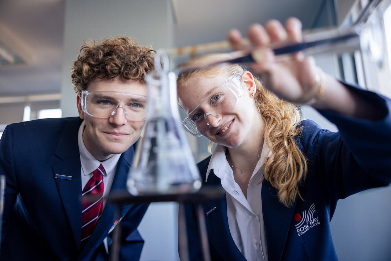 Two students in blazers and safety goggles conducting a science experiment in a school science lab. 