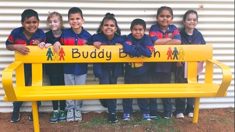 A group of 7 primary school students stand together behind a yellow Buddy Bench