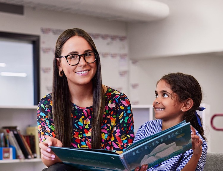 Female teacher smiles to camera with female student smiling at her as they read a book