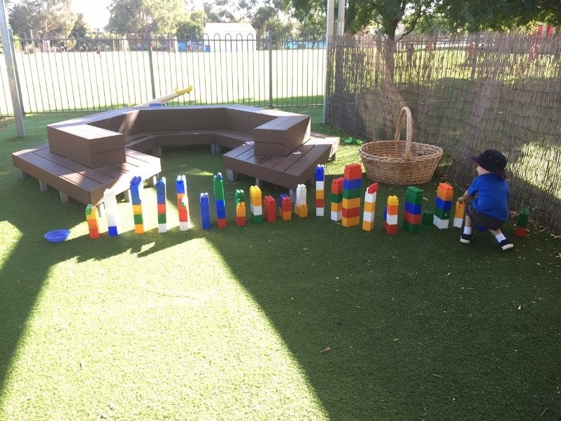 Tate kneeling next to 20 giant-lego towers of various heights. He is outside in a preschool playground on a sunny day.