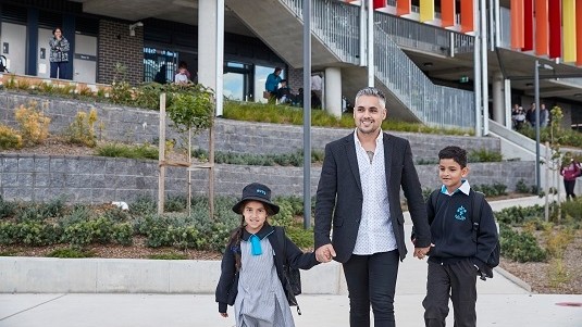 A parent or carer standing at the front of the school with their 2 children