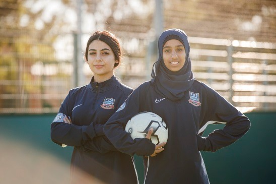 Two girls standing with outside in athletic clothing, one is holding a soccer ball.