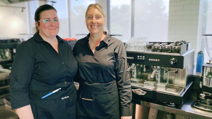 Two women standing in front of a barista machine.